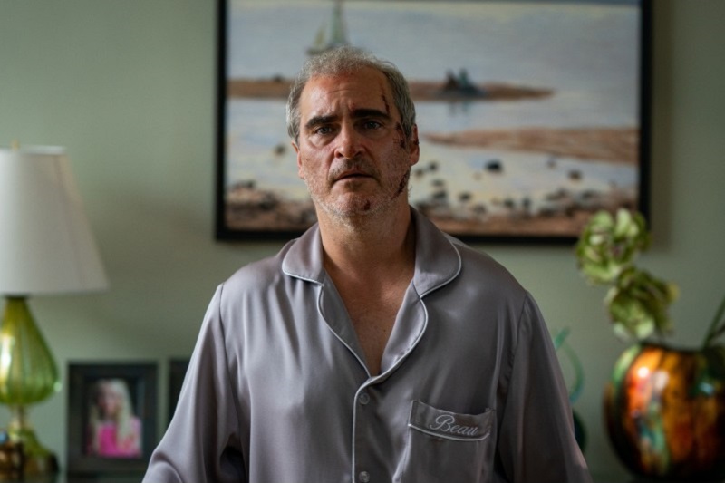 still from film 'Beau is Afraid' showing bloodied actor Joaquin Phoenix standing in a living room in pyjamas looking afraid