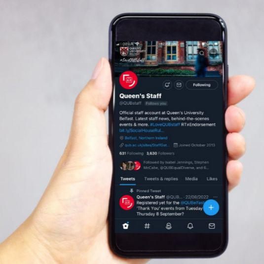 A hand holding a mobile phone with the screen showing the Queen's staff Twitter channel