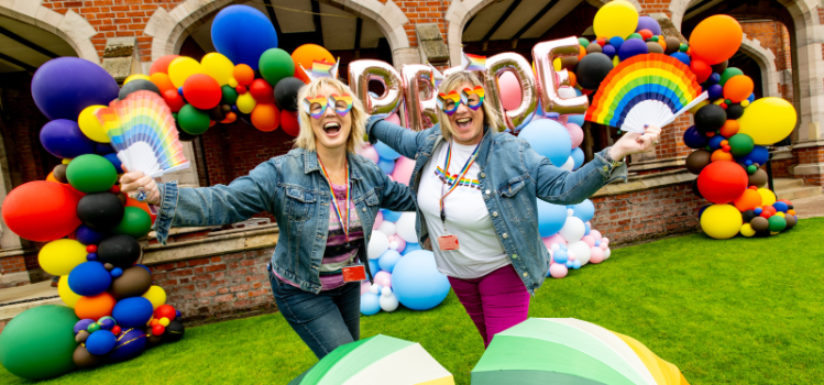 two happy staff members smiling and posing with rainbow umbrellas in front of the Pride balloon arch, Queen's University Belfast quadrangle