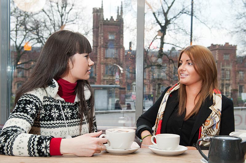 two female students drinking coffee in the students union with the Lanyon building in the background