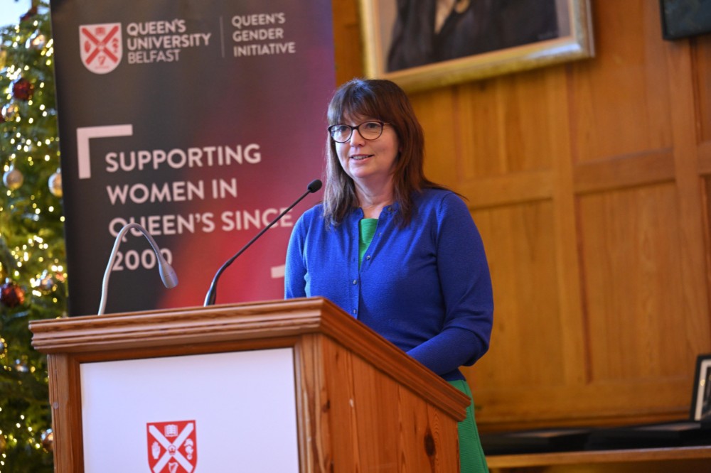 woman wearing glasses and vibrant blue top speaking from a podium. A pop-up stand in the background reads 'Queen's University Belfast / Queen's Gender Initiative / Supporting Women in Queen's since 2000'.