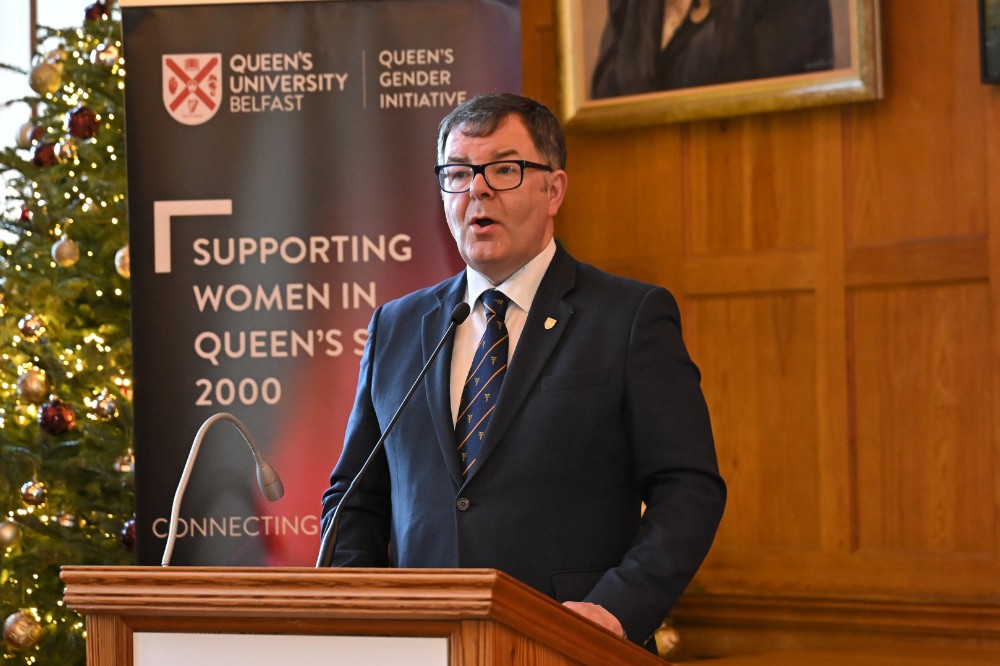 man wearing glasses and suit speaking from a podium. A pop-up stand in the background reads 'Queen's University Belfast / Queen's Gender Initiative / Supporting Women in Queen's since 2000'.