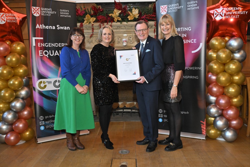 smiling woman and man, pictured with two other women, posing while holding a framed painting in an old event hall with large fireplace in the background and pop-up stands and balloon pillars on either side