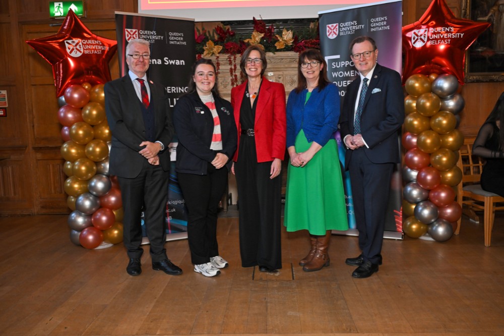group of two men and three women posing for a photo in an old wood-paneled event hall with pop-up stands and presentation screen in the background