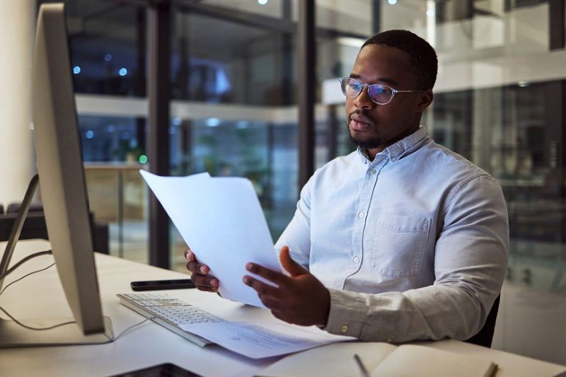 black male working late in an office, sitting at a desk with computer monitor, holding and looking at sheets of paper