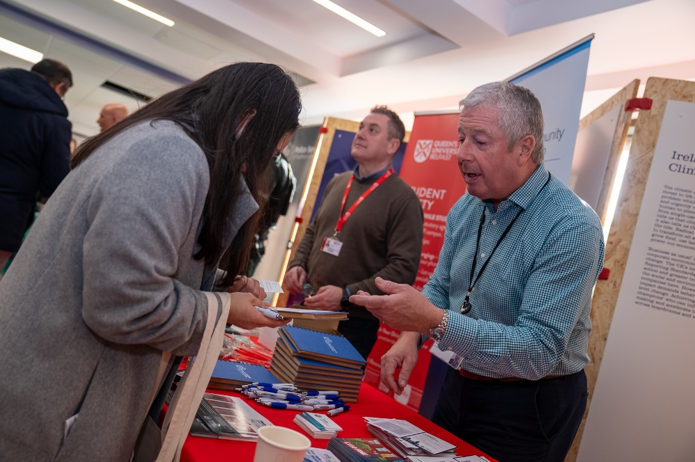 Two people talk at an indoor campus event, with one gesturing while explaining something at an information stall.