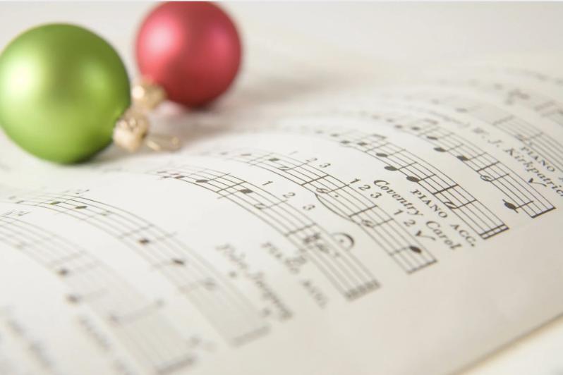 Open sheet music with holiday notes in soft focus. A red and a green Christmas ornament rest on the pages, conveying a festive, warm atmosphere.