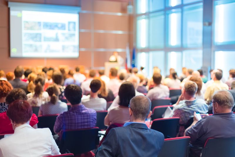 Large group of people seated at a conference with a presentation happening in the background. View is from the rear.