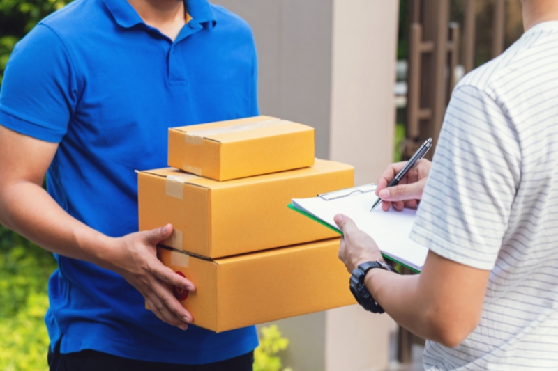 courier in blue shirt delivering three box parcels to a person holding a clipboard, pen and paper