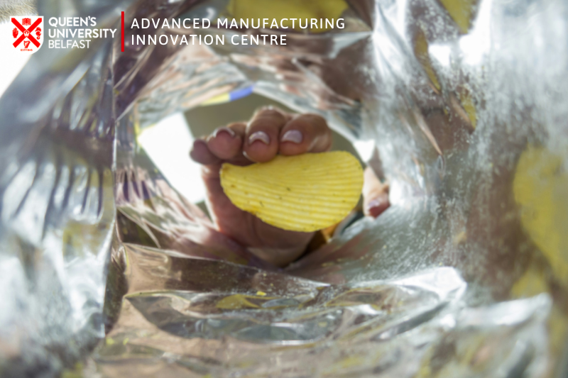 Person lifting a crisp out of a crisp packet (picture captured from inside the bag). Image includes logo of the Advanced Manufacturing Innovation Centre, Queen's University Belfast.