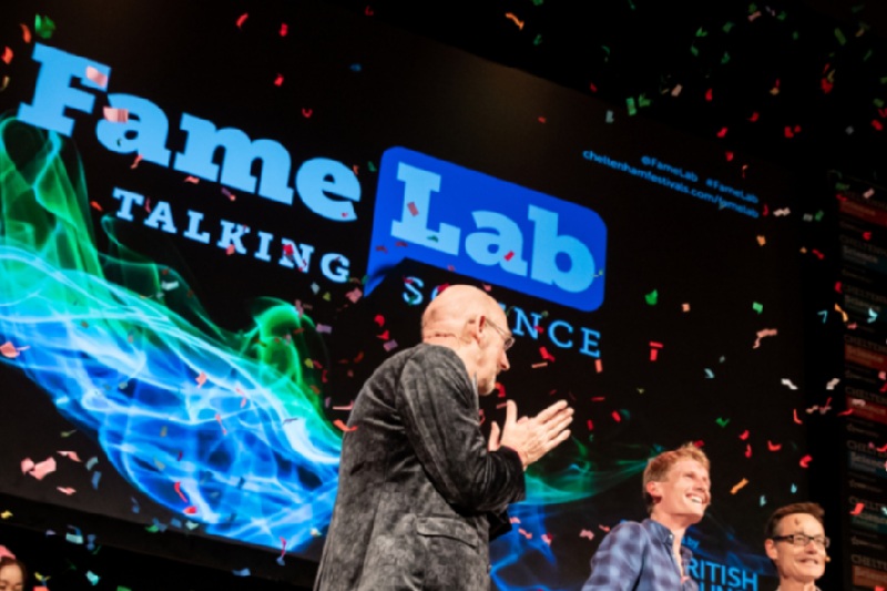 Two competition winners being applauded on stage by an emcee as confetti rains down at an event. Background presentation screen shows wording 'Fame Lab: Talking Science'.