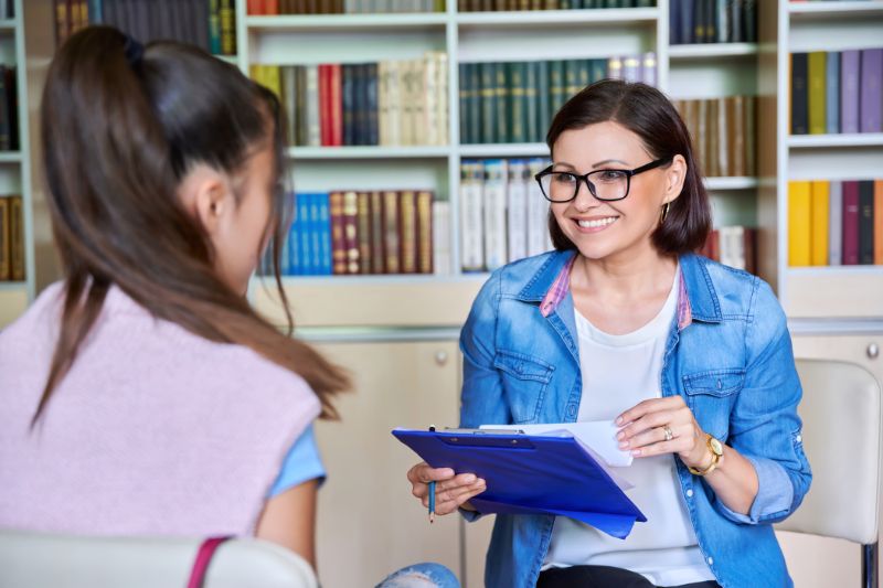 smiling woman in blue denim and glasses holding a clipboard and chatting to another young woman, with bookshelves shown in the background