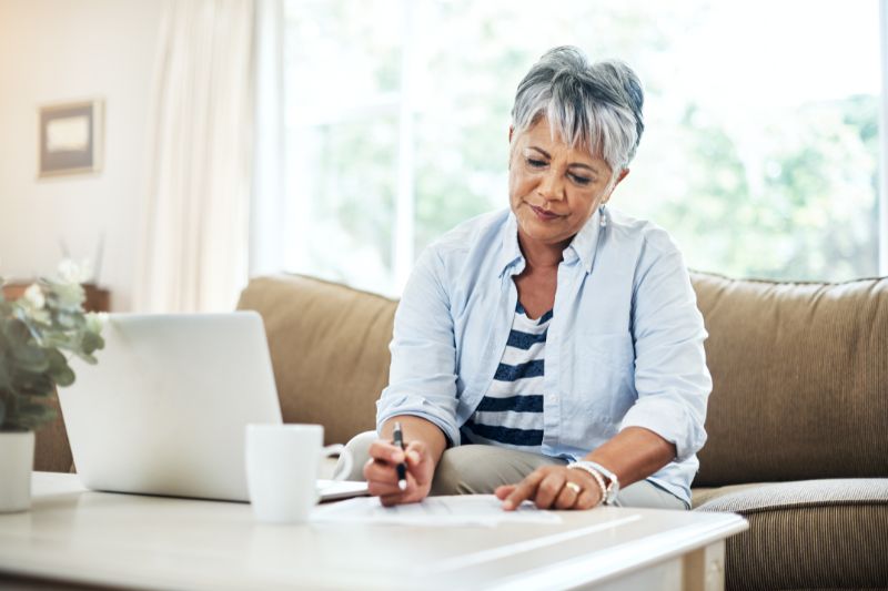 mature woman in a bright home space sat on a sofa writing and using her laptop at a coffee table