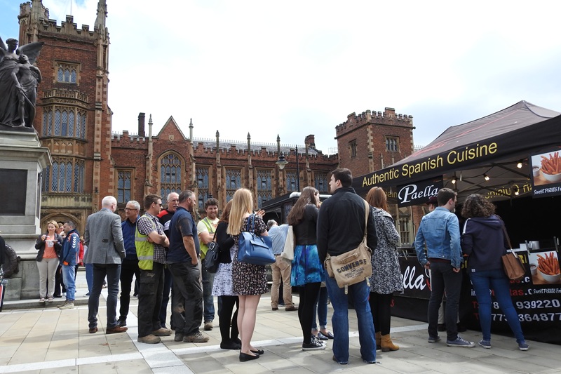 a food vending truck selling Authentic Spanish Cuisine to a large group of people in front of a university building on a sunny day