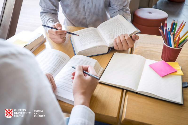 two people sitting at a desk looking at books and with pencils or colouring pencils and notepad to hand