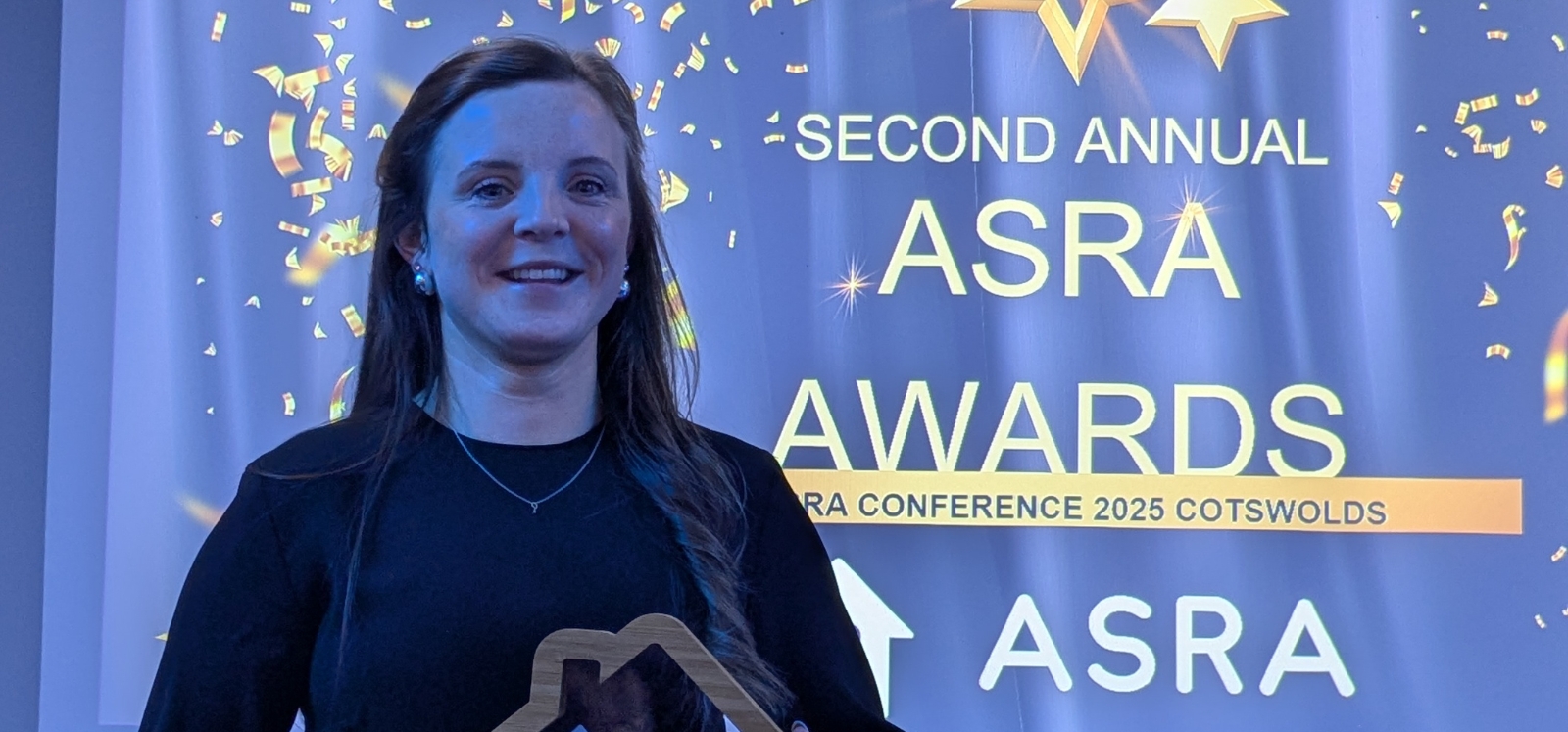 close-up of smiling woman holding an award at a gala event. Background projected graphics read
