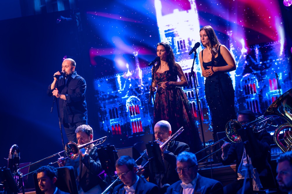 a man and two young women singing on a blue-lit stage with presentation screen behind them and orchestra in front of them in the foreground