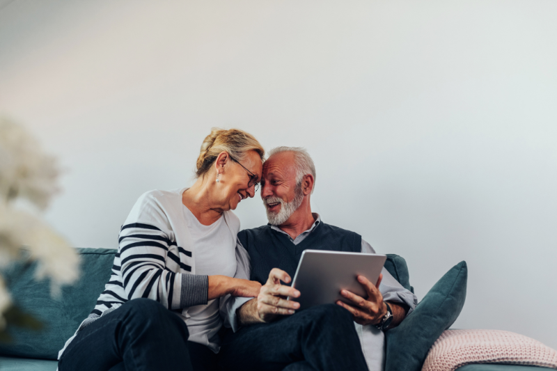 Mature man and woman, maybe a retired husband and wife, sharing a funny intimate moment on a sofa. The man is holding a tablet.