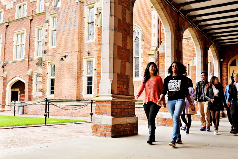 smiling, chatting group of diverse young people walking towards camera in a cloistered area of a redbrick building (Queen's University Belfast Lanyon Building)