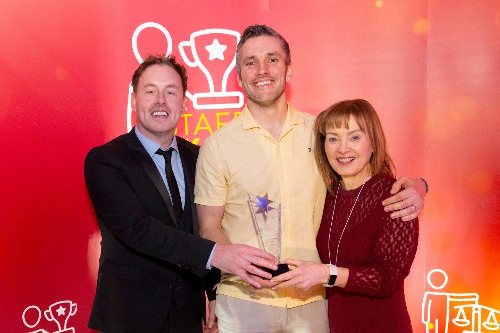 man in light-coloured shirt in centre smiling and being presented with a trophy by a man and woman at an event