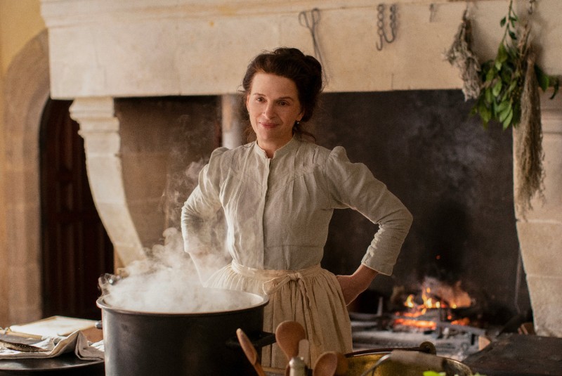 still from film 'The Taste of Things' showing smiling woman in Victorian dress standing in an old kitchen with big open fire behind and big cooking pot in front