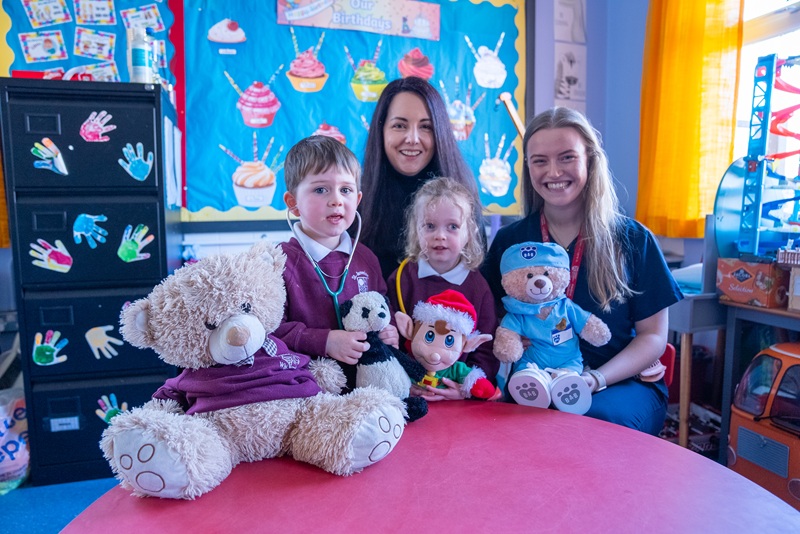 two young adult females and two nursery age children holding teddy bears in a nursery school classroom or childcare setting