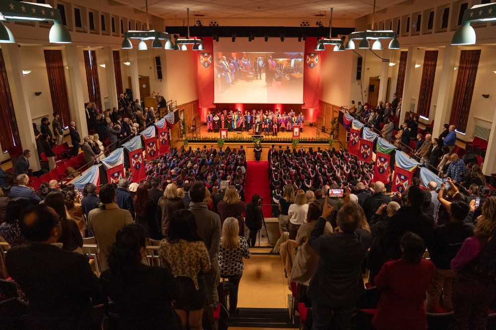 large balconied hall with many people standing upstairs in the foreground and on the ground floor at a graduation event