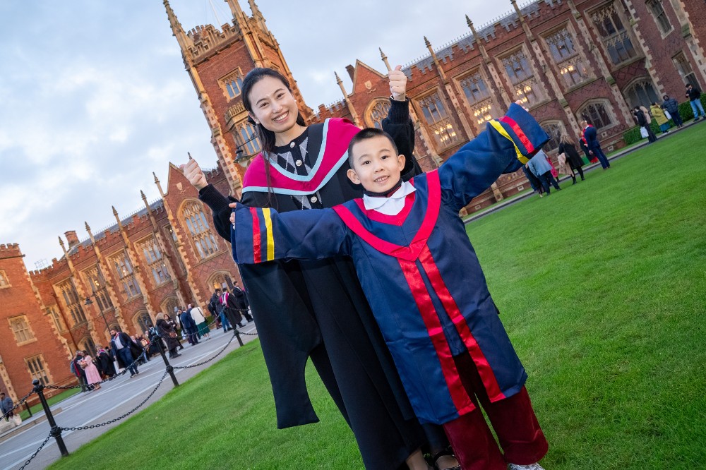 young woman of Asian origin celebrating her graduation with a young boy on the lawn of an old redbrick building